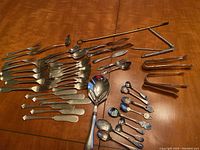 Full overview of silver plated flatware including knives, forks, spoons, and metal tongs spread on wooden table.