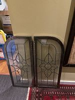 Two arched stained glass window panels leaning against a wall, showing their geometric floral leaded glass design, black metal frames, and clear textured glass.