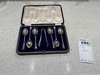 Boxed set of six silver tea spoons and one pair of silver sugar tongs arranged in blue velvet-lined box with white lid lining.
