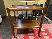 Two-tier wooden tea trolley on caster wheels shown from front angle on a red patterned carpet with bar stools and kitchen appliances in background.
