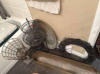 Photo showing multiple black wire planters, brass metal tray, grapevine wreath, and brass fish kettle arranged on floor near wall corner.