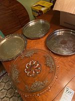 Four silver plated trays with different etched floral designs and raised beaded edges, placed on wooden table.