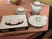 White ceramic tea pot, coffee pot, boxed 3 piece server, and covered casserole on table with pink cloth