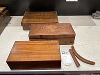 Three wooden boxes placed on a countertop, two boxes show engraved initials on top surfaces. Two curved wooden pieces are beside the boxes.