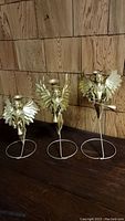 Three gold-tone metal angel candle holders standing on a dark wooden surface in front of wooden panel background, seen from front angle.
