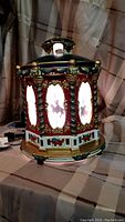 Hexagonal ceramic lighted carousel with illuminated silhouette scenes of Christmas figures, holiday decor with red, green, white, gold accents.