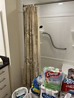 Overview of bathroom corner showing beige patterned shower curtain and stacked packages of Charmin and Royale toilet paper, paper towels, towels, batteries, and bathroom supplies on floor.