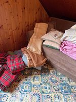 Four red tartan fleece blankets, beige and brown knit blankets stacked near couch on patterned carpet and wood panel wall