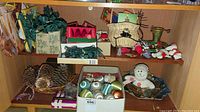 Full shelf view showing assorted Christmas ornaments including boxed glass balls, pinecones in baskets, snowman figure, and string lights piled within boxes and bags.