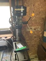 Front and side view of black Mastercraft drill press with yellow handles, mounted on a metal base attached to workbench in workshop.