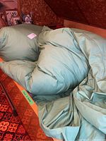 First image shows one of the twin eiderdown duvets with a green duvet cover, wrinkled and resting on a bed with a patterned red and brown carpet visible.
