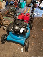 Front view of blue Yard Works gas lawn mower with two red gas cans on the deck in garage environment.