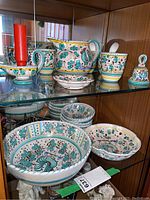 Display of the pottery set including jug, cups, bell, candle holder, and spoon dish with colorful rooster and floral patterns.