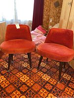 Pair of orange crushed velvet upholstered slipper chairs with curved seat and wood legs with brass feet caps on a patterned carpet.