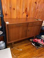 Full view of mid century buffet/server showing two drawers above two lockable doors on tapered legs, wood finish with wood panel wall behind.