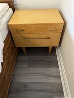 Full view of the wooden night stand placed next to a bed, showing the two drawers and the drop-front compartment closed.