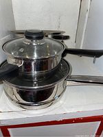 Two stacked stainless steel pots with black handles, one with glass lid, placed on a white shelf.