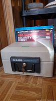 Front view of beige Chubb fire-safe security chest with original box in background showing "Chubb On Guard Fire-Safe Security Chest" branding.