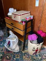 Wooden sewing cabinet with drawers and shelf, small woven sewing basket on top, bags of yarn and sewing supplies around it.