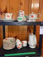 Partial set of ivory-colored dishes with orange floral pattern on black shelving unit against wooden wall. Includes plates, cups, coffee pot, tea pot, creamer, and platter.