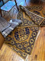 Photo showing two yellow and blue wool rugs partly rolled and laid flat on hardwood floor, one larger than the other, with two grey woven mats rolled up beside them.