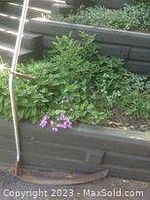 Photo of an antique scythe lying next to a raised garden bed with plants and flowers. The scythe features a metal curved blade and a long wooden handle in gently used, antique condition.