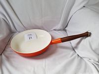 Orange enamel cast iron fry pan with white interior and wooden handle placed on fabric background.