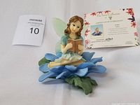 Front view of the Dezine collectible fairy figurine seated on a blue flower, with certificate and auction tag in the background.