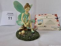 Front and angled view of the light green fairy figurine seated on a mushroom base, showing detailed wings, dress, and base. Certificate of Authenticity displayed next to it.