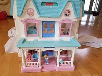 Front view of the doll house displaying the porch, blue doors, windows with pink frames, three figurines including a baby on a porch swing, and the pastel colored house roof.
