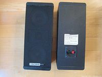 Front and back view of the pair of Koss Dynamite M/85 bookshelf speakers showing the speakers with front black grilles and the rear panel with model label and wire connectors.