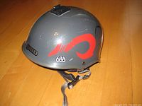 Side view of gray Giro 686 helmet showing red 686 logo and stylized animal head graphic, with chin strap lying on wooden floor.
