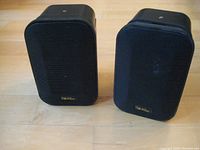 Front view of the pair of black AR-225 weatherproof speakers placed on a wooden floor showing the metal grille and AR label.