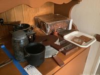 Wide angled photo showing antique hinges mounted on wooden plate, Mason jar, black grinder, wood bellows, box with metal hinges, rectangular metal plate, and vintage photo frame with old photo inside on tray.