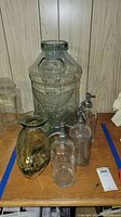 Photo showing six glass items on wooden shelf near wall, including large clear glass jug with embossed eagle, amber vase, two siphon bottles with metal tops, and two clear bottles without tops.