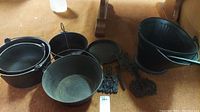 Three cast iron pots, one metal pot (coal scuttle), two metal decorative wall mounts, cast iron frying pan, and metal shovel arranged on a floor.
