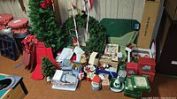 Wide view of Christmas decorations including artificial trees, wreaths, ornaments, boxes, and holiday items spread on the floor against a panel wall.
