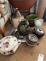 Overview photo showing large corked jug, floral lidded pot, green planter, chamber pot, napkin rings on tray, and glass vase on carpeted floor
