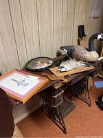 Wood-like goose decoy, fish plaque, metal trays, and framed goose print displayed on wooden furniture against wood-paneled wall.