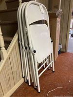 Four white metal folding chairs stacked together, showing front and side angles. Scratches and wear visible on seats, backrests, and frames. Chairs lean against a wall near stairs indoors.