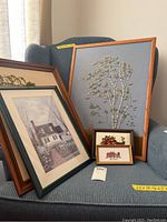 Photo showing six framed art pieces on a chair, including embroidery and print, with measuring tape for size reference