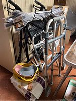 Photo showing two folding metal walkers alongside a walker with seat, brakes, and basket. Multiple bedpans in yellow and grey are stacked on the floor.