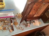 Photo of assorted costume jewelry and two Timex wristwatches on a wooden surface near a jewelry cabinet including gold-tone necklaces, bracelets, and a pearl necklace.