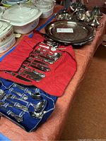 Photo of trays, flatware, and some serving pieces on table with red and blue fabric rolls holding silver flatware sets. Several serving spoons and ladles visible along with large metal tray and sugar and creamer set.