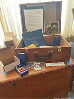 Open wooden box showing an array of military memorabilia including patches, pins, certificates, and papers inside and around with a handle and brass latches visible.