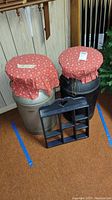 Two metal milk cans with red floral cushions on top and small black wooden shelf in front.