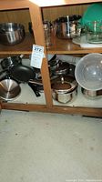 Cabinet shelf with stainless steel pots, pans, and lids stacked and arranged, some nonstick visible, under shelf has clear plastic bowl and other cookware
