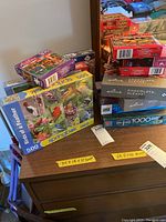 Full lot of assorted jigsaw puzzle boxes stacked on a small table, featuring different brands, piece counts, and themes