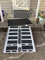 Folding aluminum ramp unfolded and placed on concrete steps leading to a door. Black non-slip strips visible on the surface.