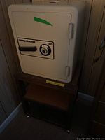 White Sentry Safe with combination lock and handle sitting on a wooden rolling cart with caster wheels, dark brown frame and a middle lighter colored shelf.